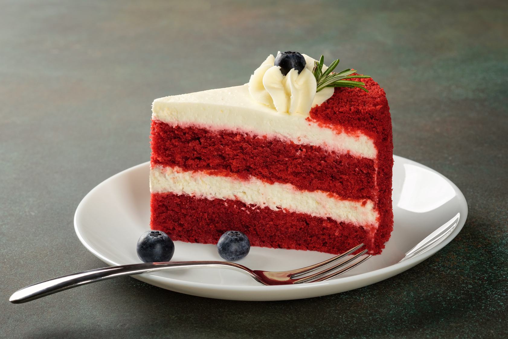 Red Velvet cake with blueberries on white plate over dark green background.